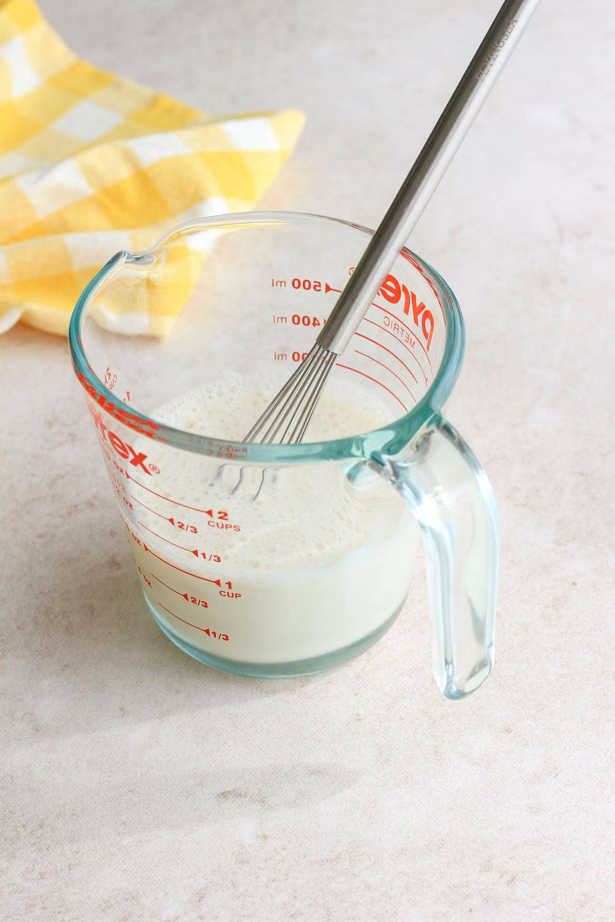 From the side, a liquid measuring cup with a mini whisk after mixing soy milk with vinegar.