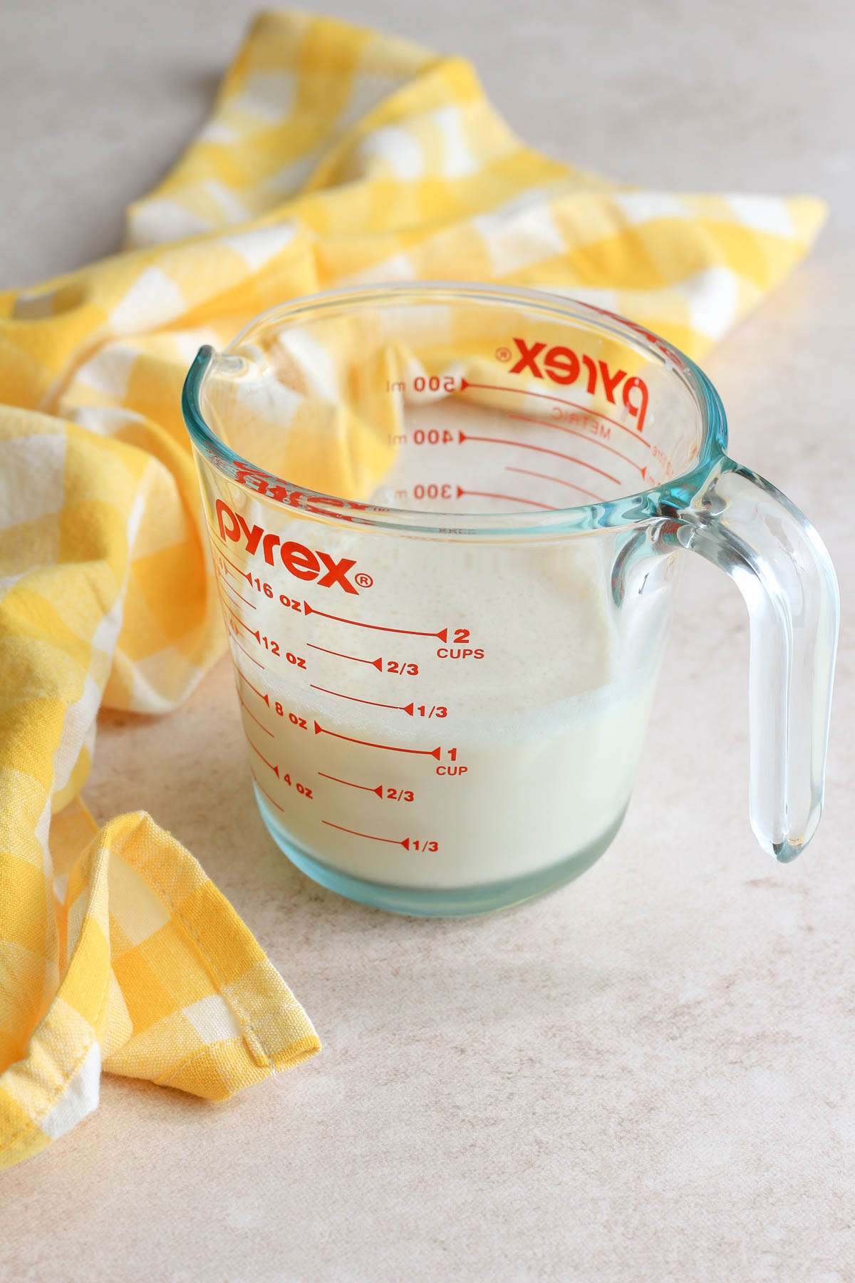 A liquid measuring cup with homemade vegan buttermilk next to a yellow buffalo check towel.