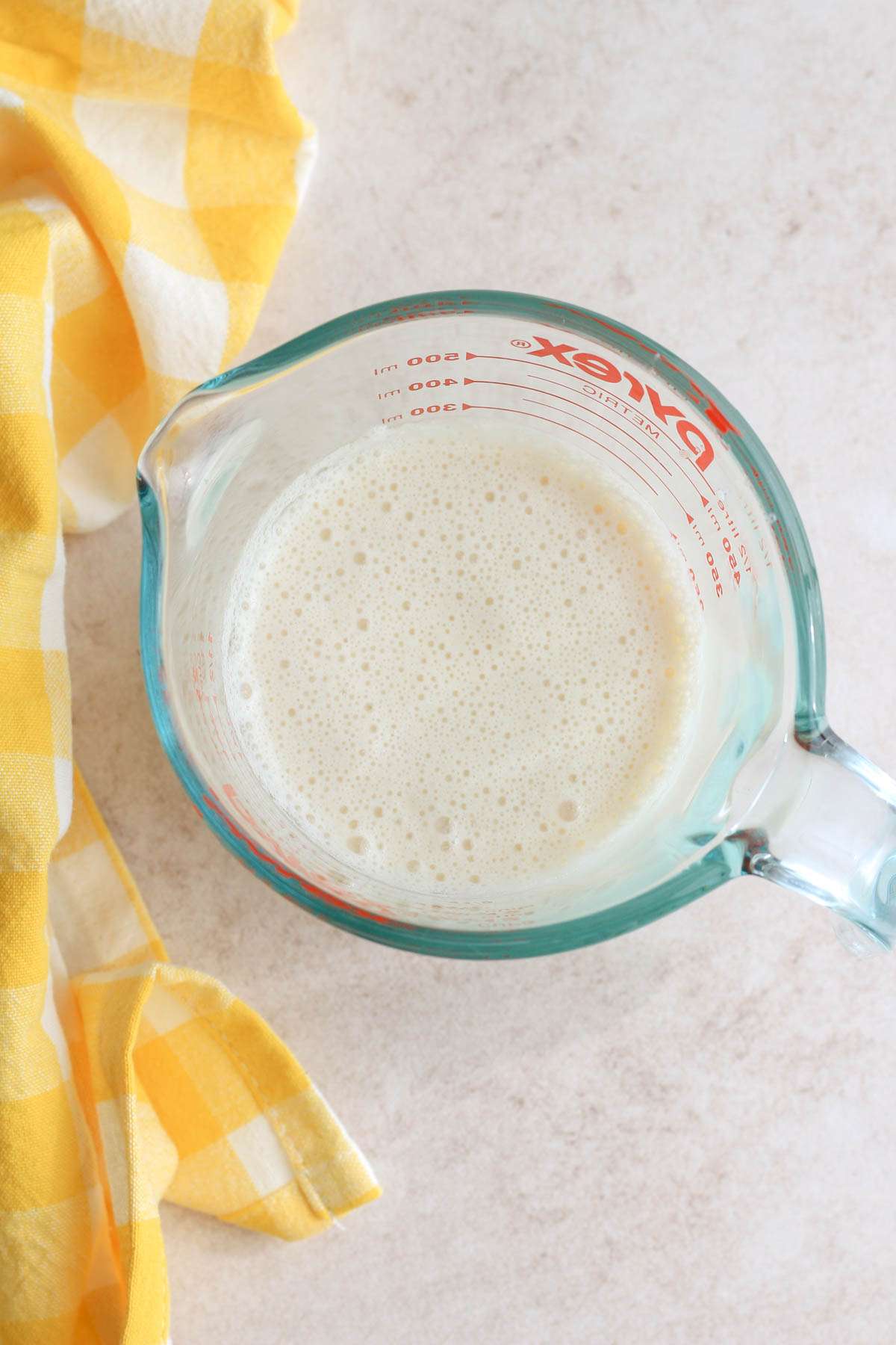 Top down image of homemade vegan buttermilk with soy milk in a liquid measuring cup next to a yellow an white buffalo check towel.