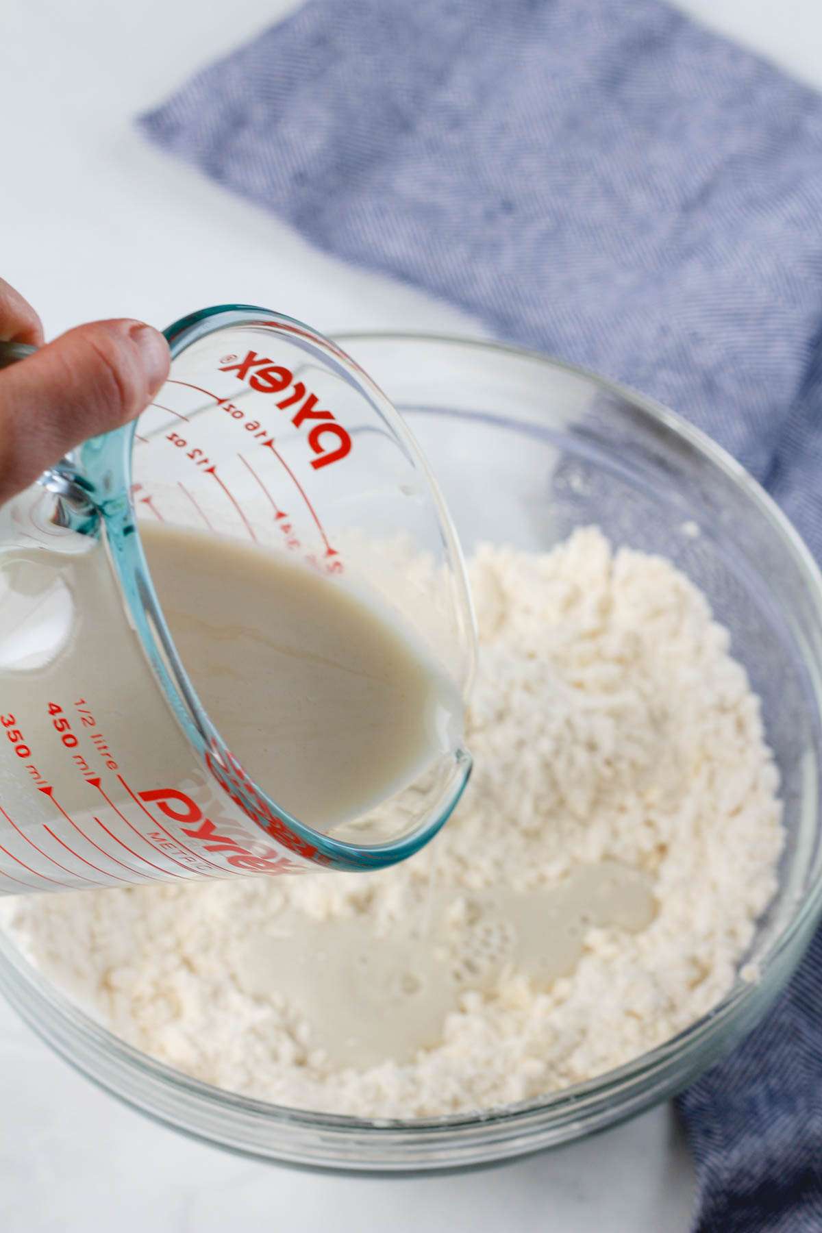 A liquid measuring cup pouring dairy-free buttermilk into a bowl of biscuit dough.