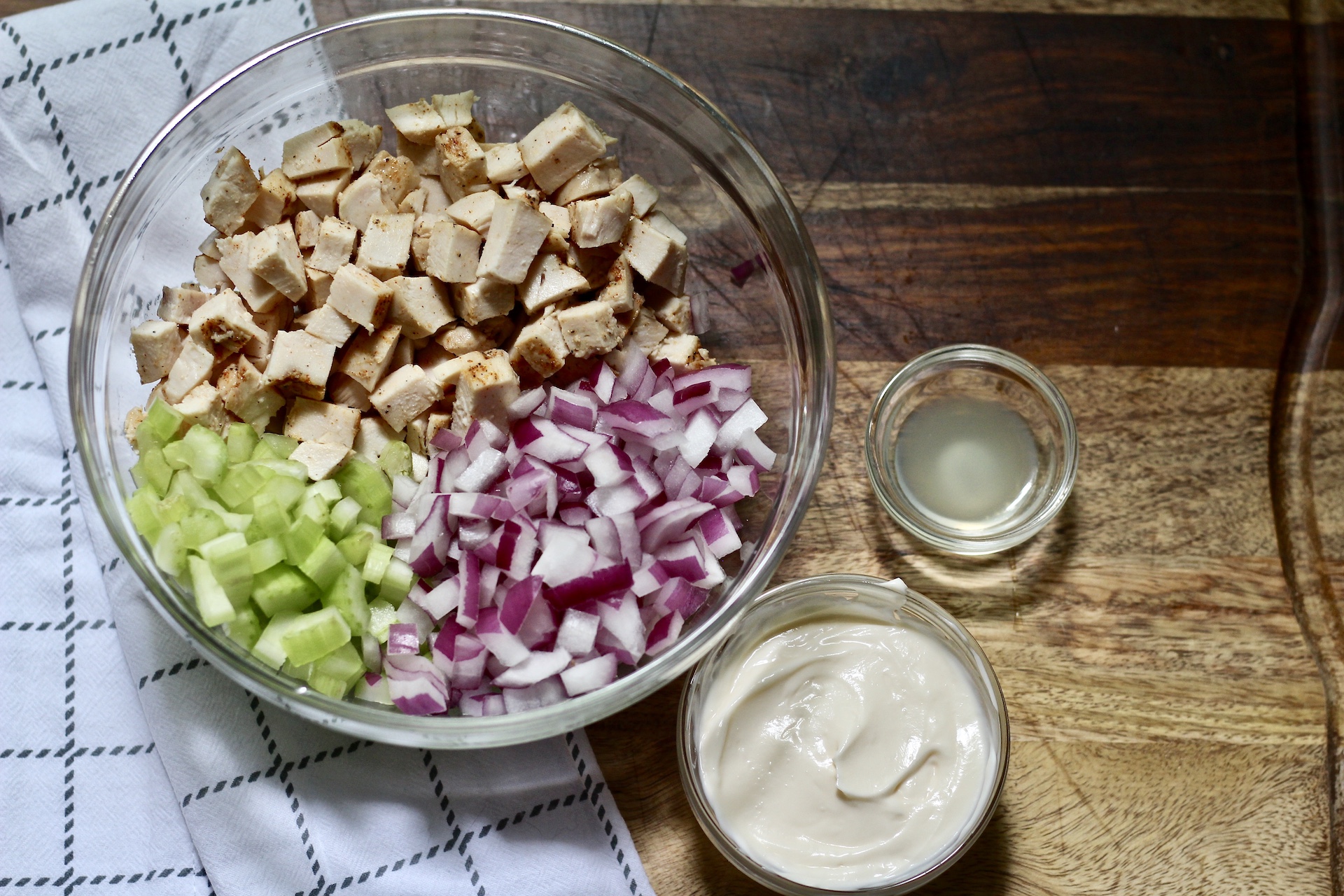 Bowl of chicken, celery, and red onion with a bowl of mayo and lemon juice on a cutting board.