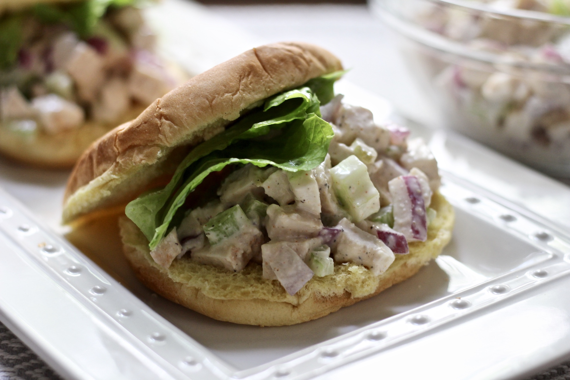Chicken salad, on a bun, on a white platter, with a bowl of chicken salad in the back. 