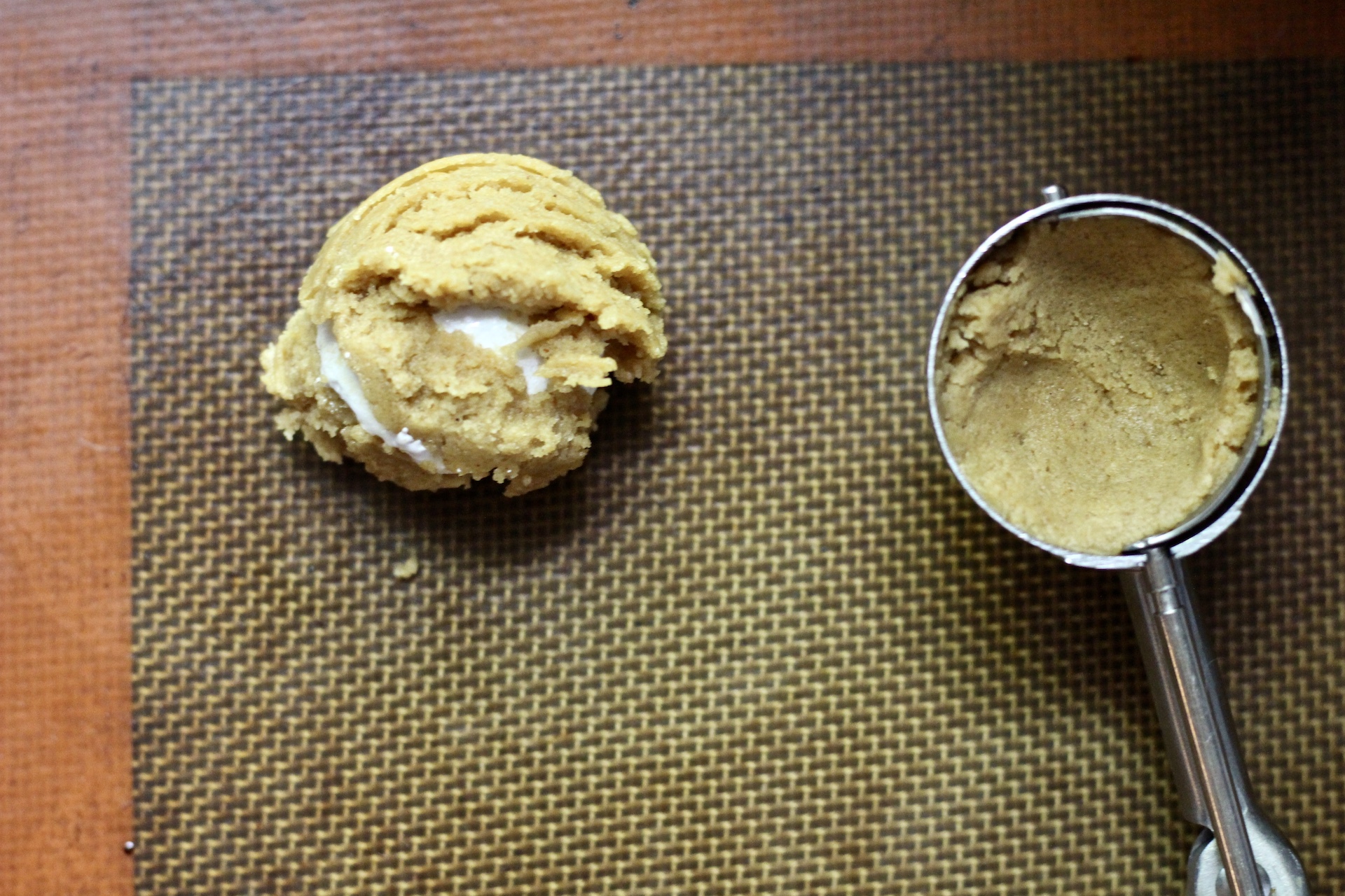 A scoop of fluffernutter cookie dough next to a cookie scoop with peanut butter cookie dough on a nonstick backing mat.