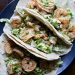 Three shrimp tacos on a wooden cutting board with a dark gray plate.