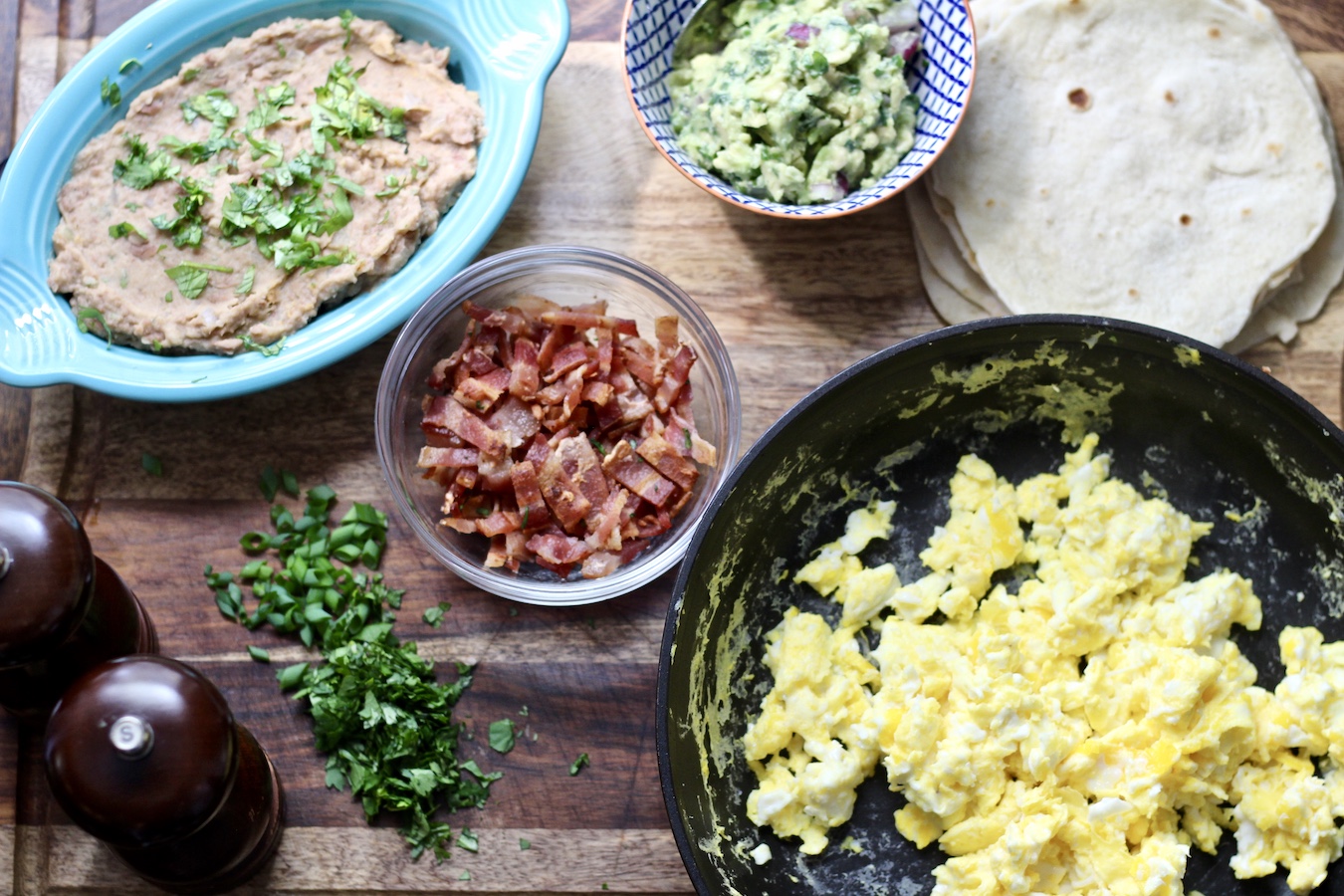 The ingredients for breakfast tacos, scrambled eggs, bacon, refried beans, guacamole, and cilantro.