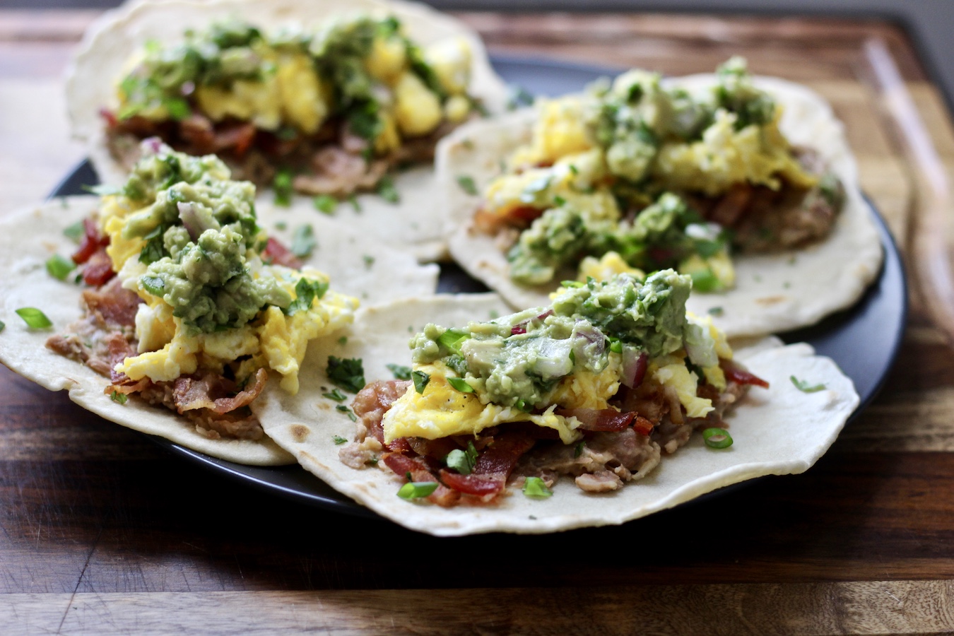 Four flat breakfast tacos on a grey plate sitting on a wooden cutting board.