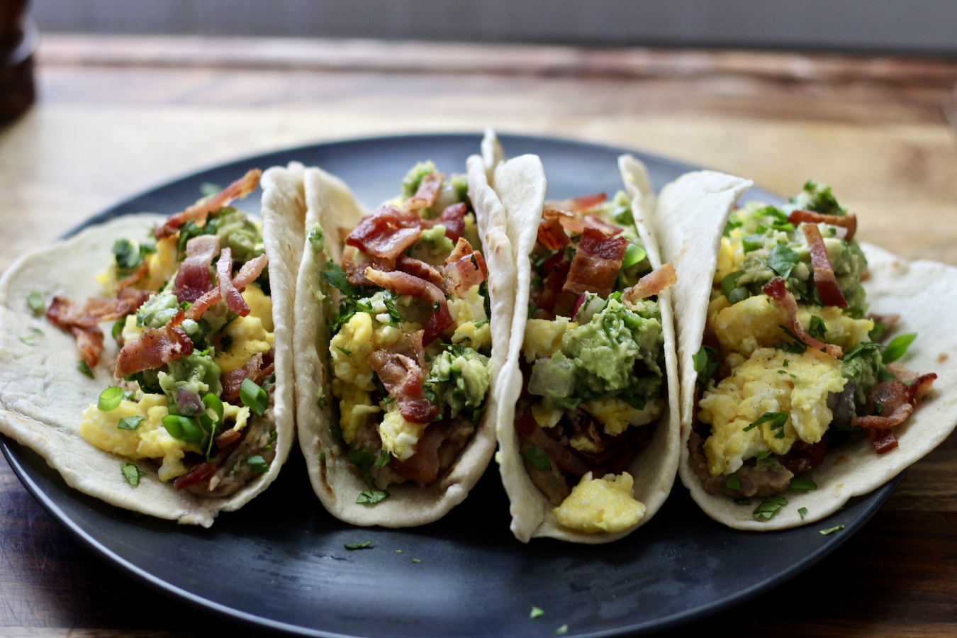 Four breakfast tacos on a dark grey plate on a wooden cutting board.