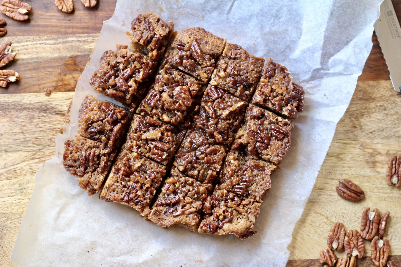 Pecan Pie Blondies on a parchment paper on a wooden cutting board with pecans scattered around.