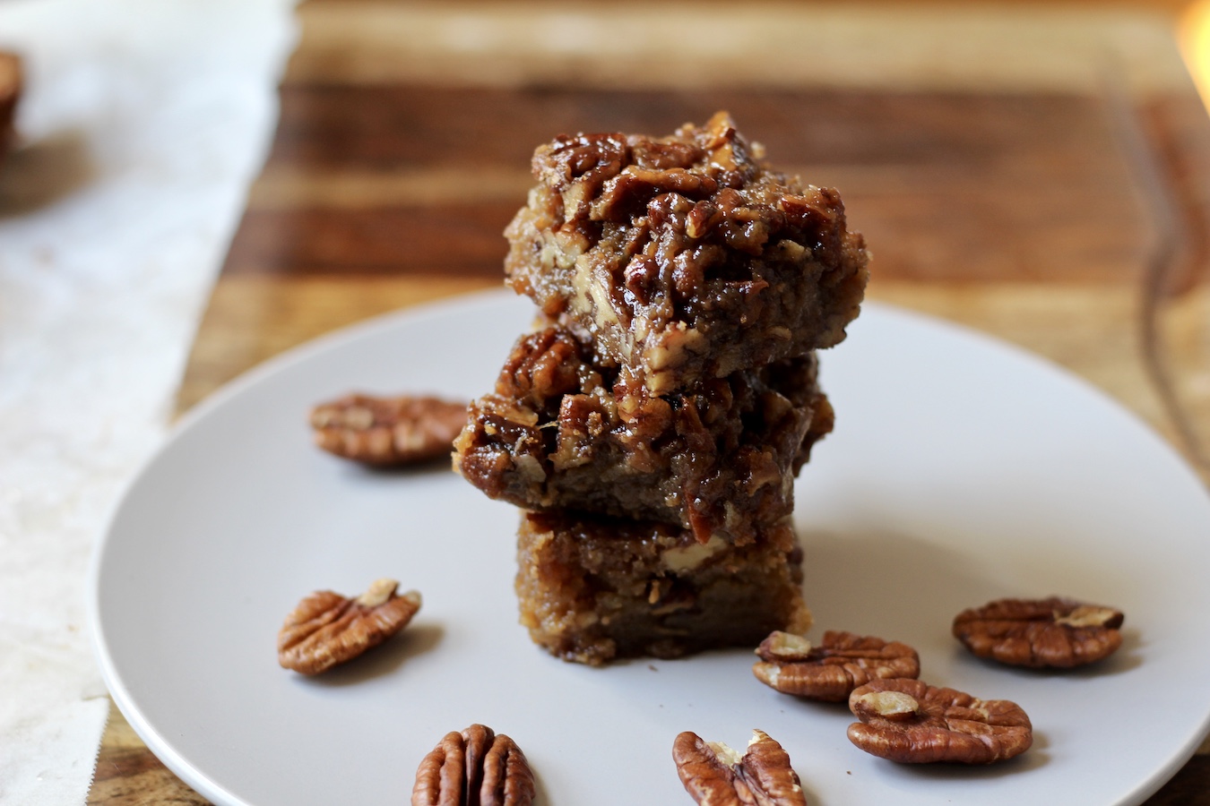 A stack of three Pecan Pie Blondies on a plate on a wooden base with pecans scattered around.