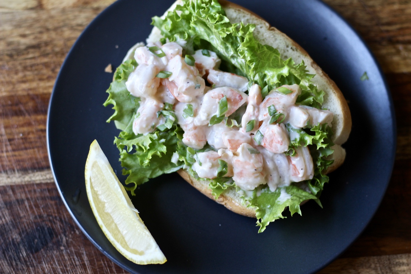 A shrimp roll on a black plate with a lemon wedge on a cutting board.