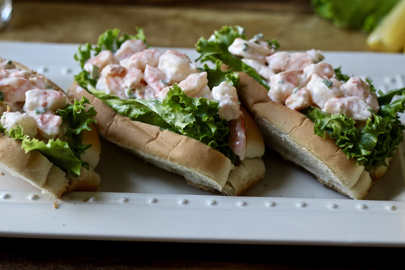 Three shrimp rolls on a white platter on a wooden surface with a lemon in the back.