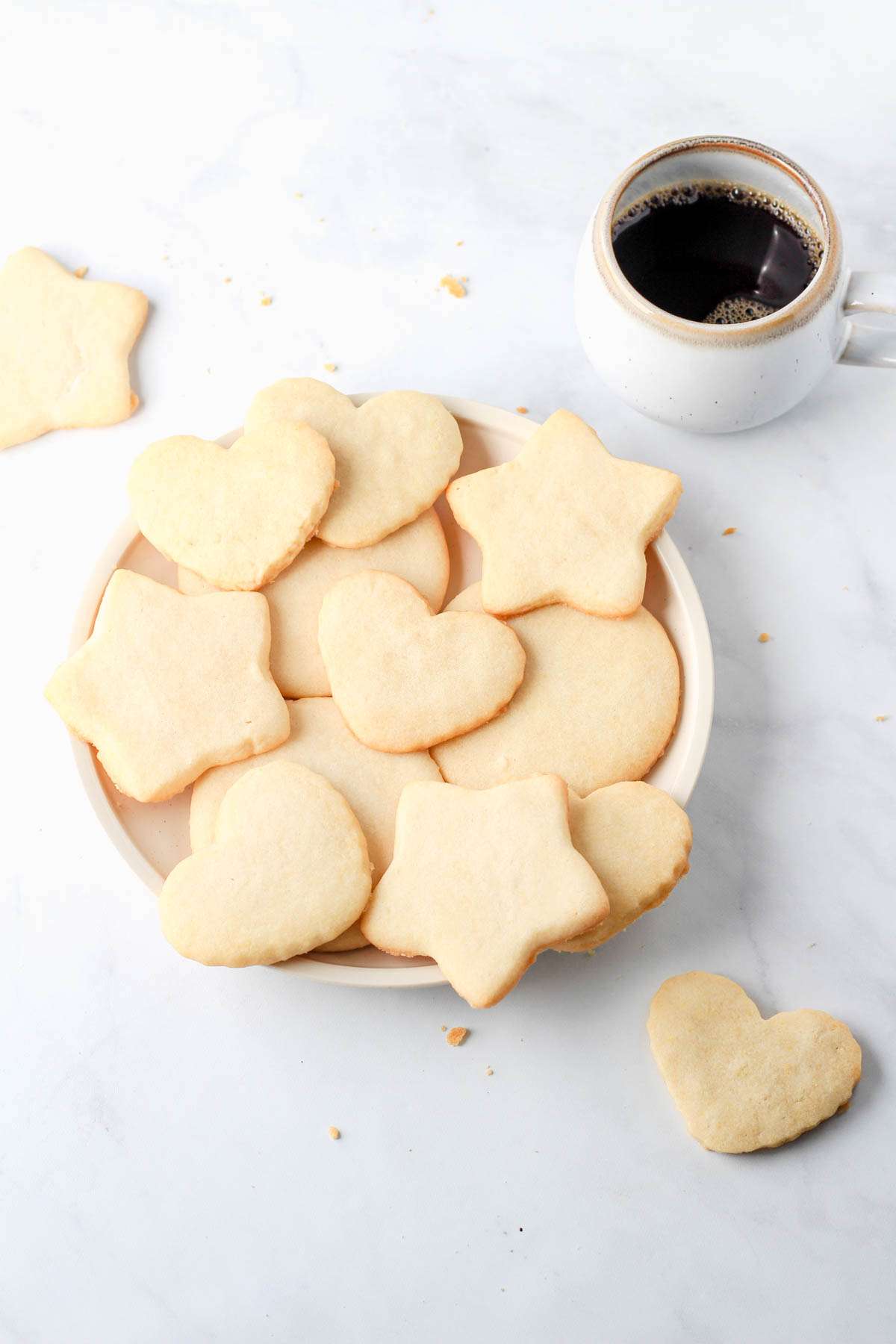 A white plate on a white counter with heart, star, and circle shaped dairy-free sugar cookies with a cup of coffee in the top right.