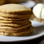 A stack of 5 pumpkin spice pancakes on a white plate with a silver fork on the right of the plate and pumpkins in the back.