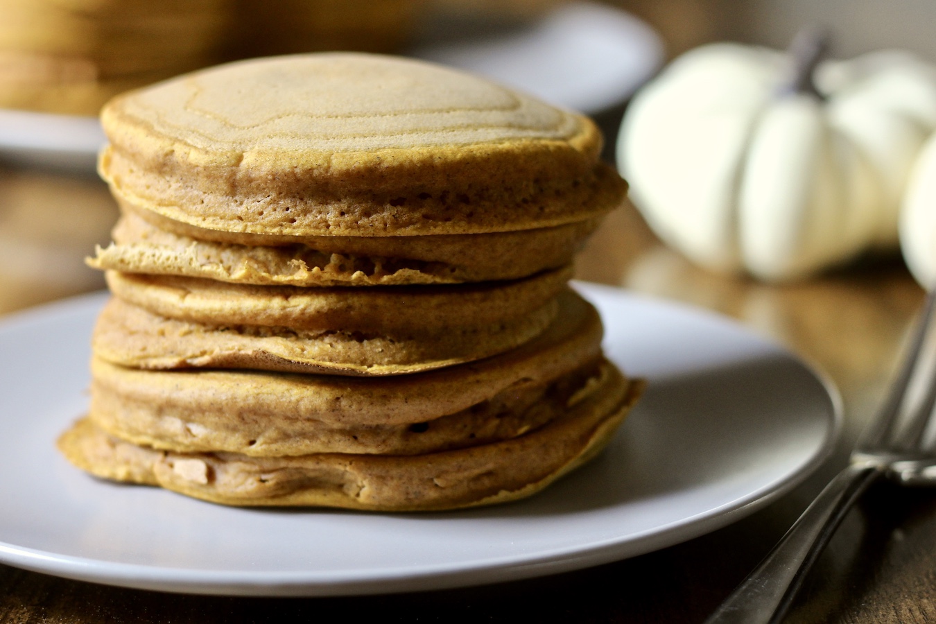 A stack of 5 pumpkin spice pancakes on a white plate with a silver fork on the right of the plate and pumpkins in the back.