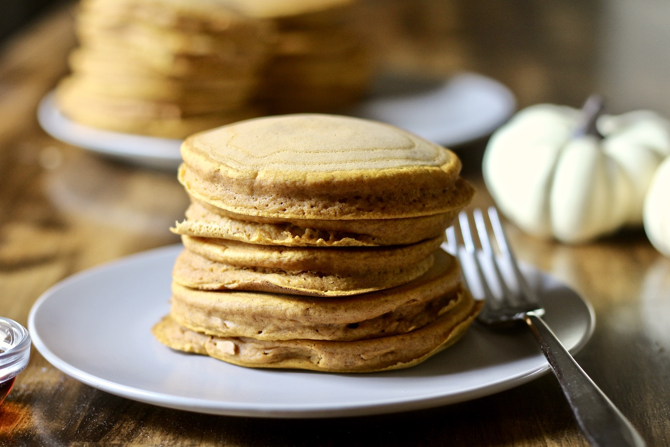 A stack of 5 pumpkin spice pancakes on a white plate with a silver fork on the right and a small bowl of maple syrup on the left. Two white pumpkins and a stack of pancakes in the back.