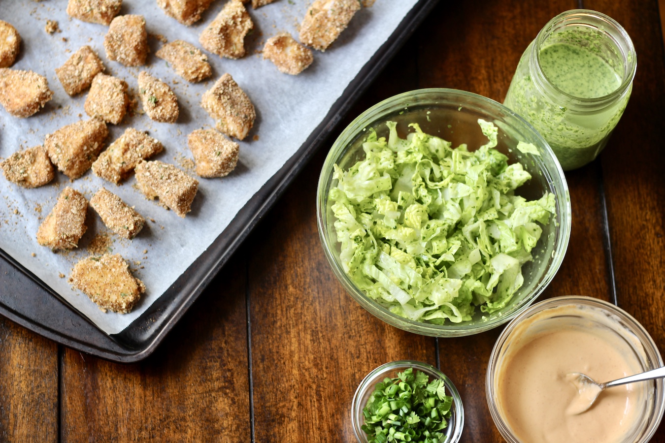 A photo of the ingredients used in fish tacos. Fish pieces on a baking sheet to the left with cilantro lime dressing, lettuce, cilantro, green onion, and a spicy mayo dressing.