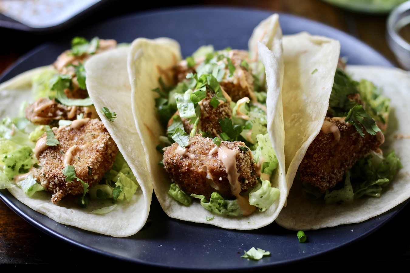 Forward looking picture on three fish tacos in flour tortillas dressed with cilantro, green onion, and dressing on a blue plate.