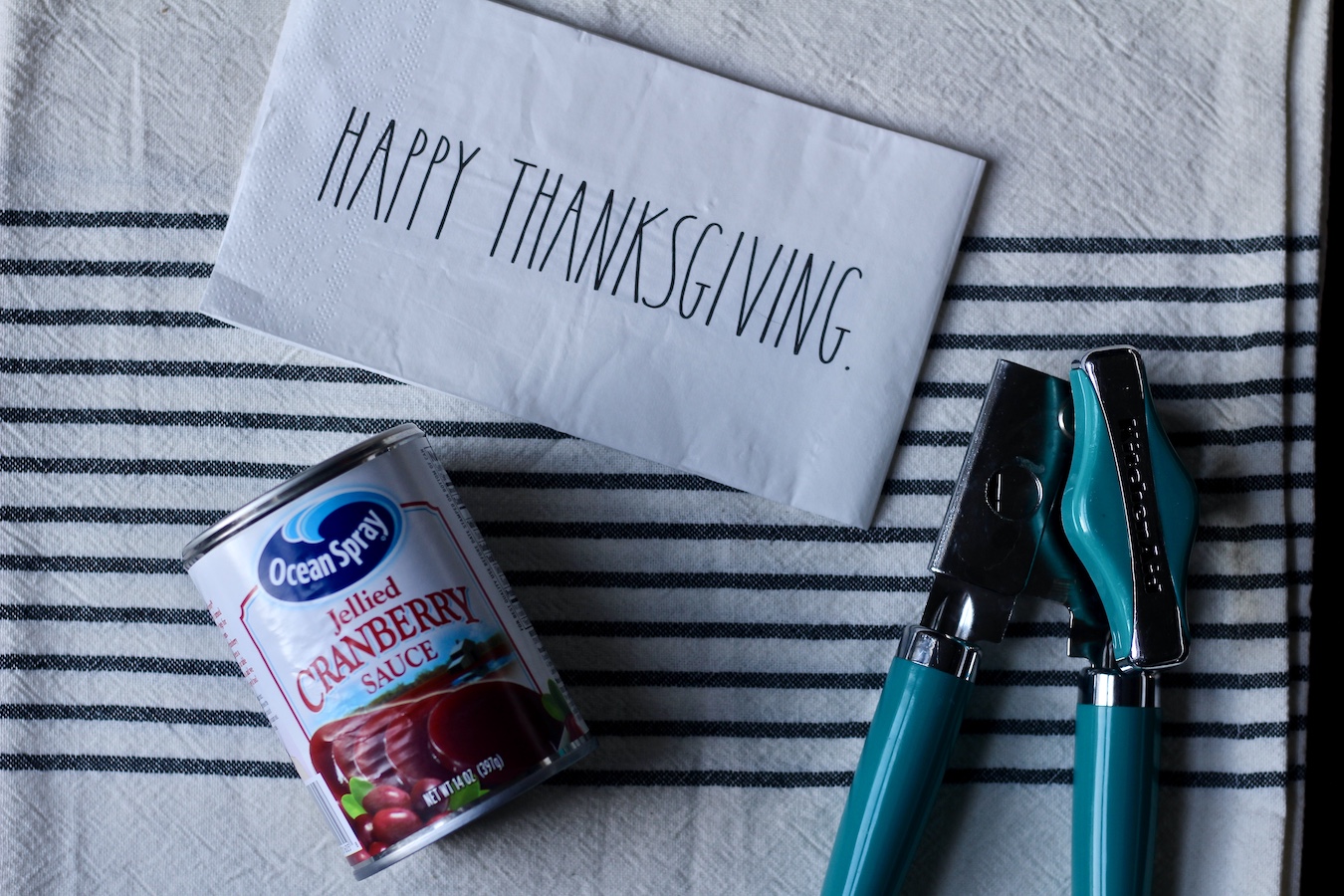 A can of cranberry sauce on a white and blue dish towel with a can opener to the right and the words happy thanksgiving on the top.
