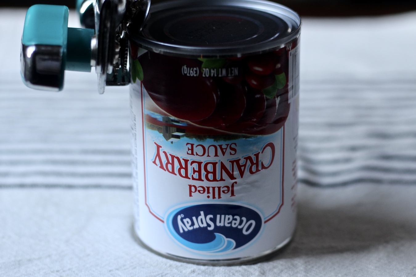 A can of cranberry sauce with a can opener on the top on a white and blue dish towel.