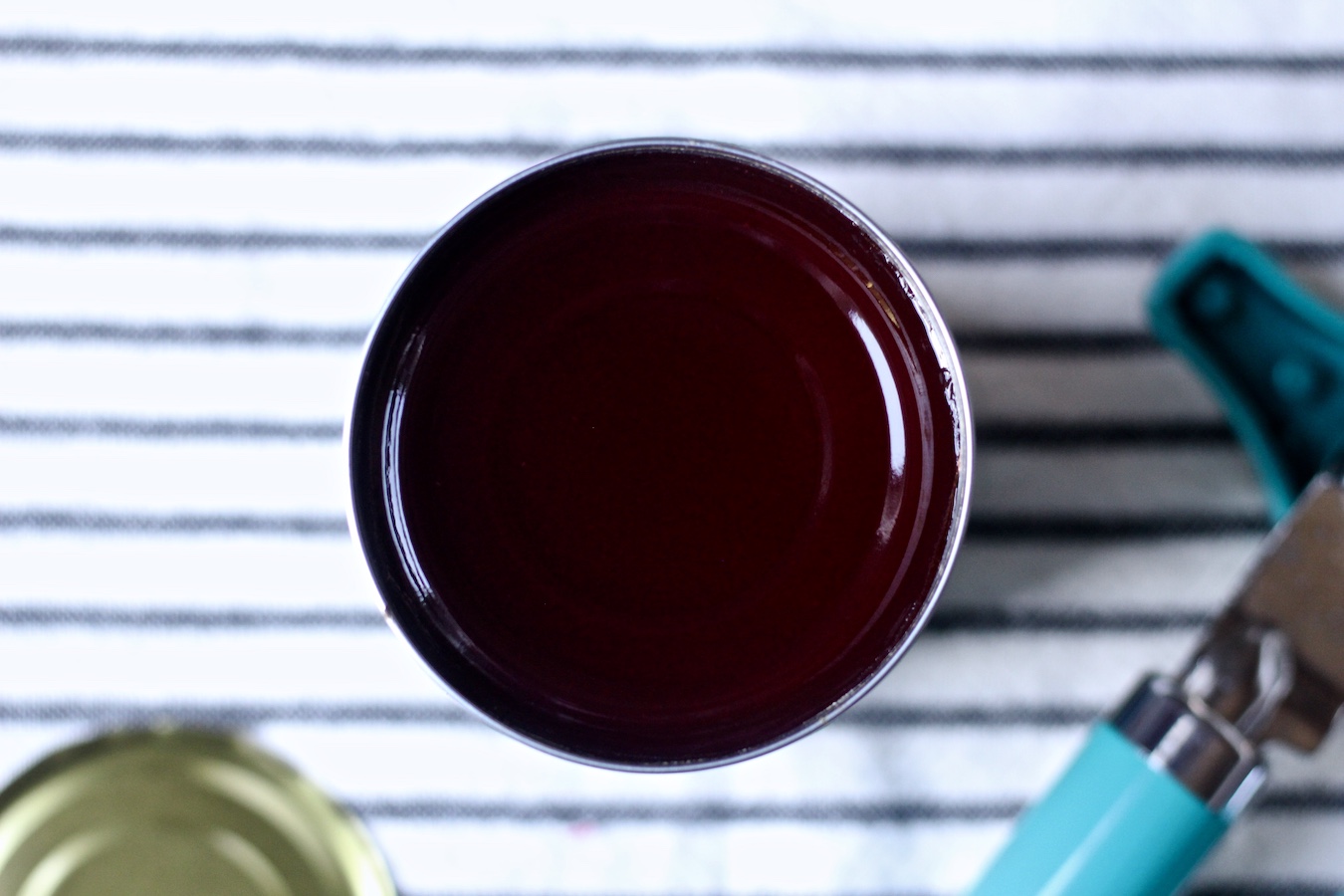 An open can of cranberry sauce with a can opener and white and blue dish towel.