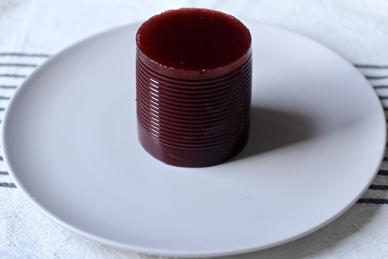A tall can of cranberry sauce on a white plate on a blue and white striped dish towel.