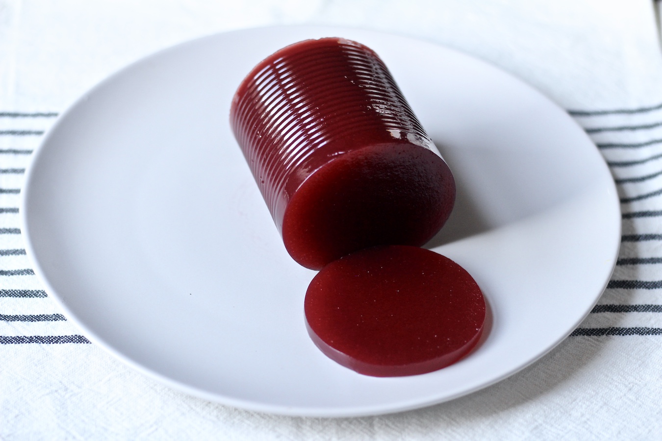 A can shape of cranberry sauce with one slice of cranberry on a white plate lofted on a blue and white striped towel.