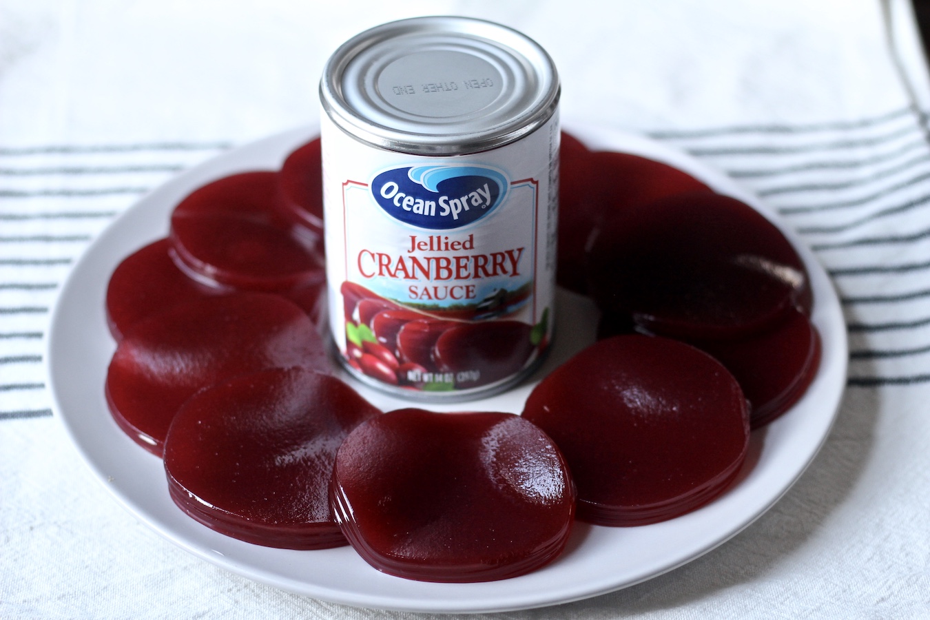 A white plate of sliced cranberry sauce with an Ocean Spray can in the middle on a white and blue striped dish towel.