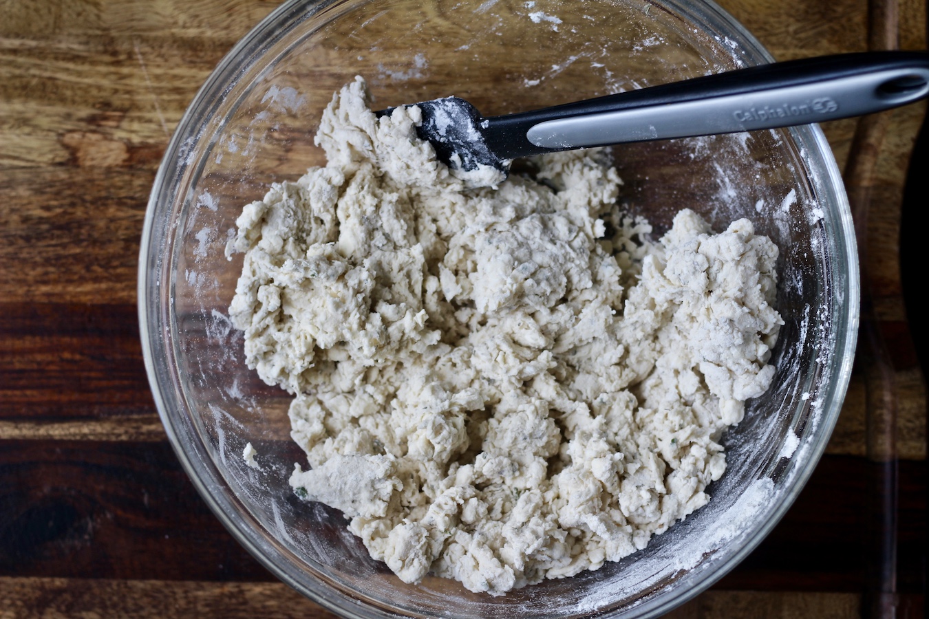 Glass bowl with a black rubber spatula. Garlic and herb biscuit dough in the bowl on a wooden cutting board.