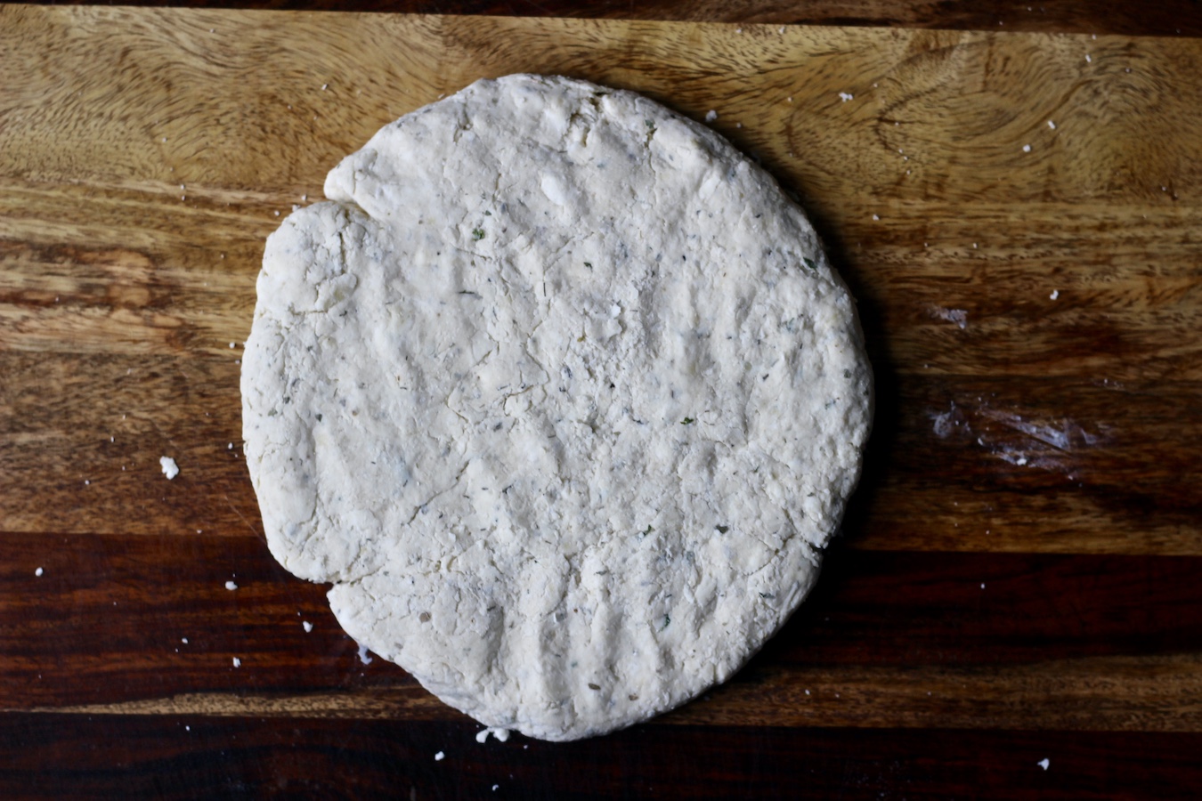A round disk of garlic and herb biscuit dough on a wooden cutting board.
