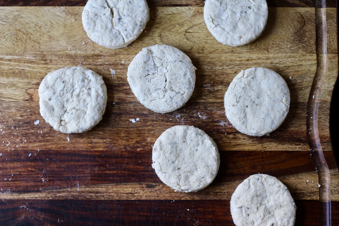 Seven uncooked garlic and herb biscuit doughs on a wooden cutting board.