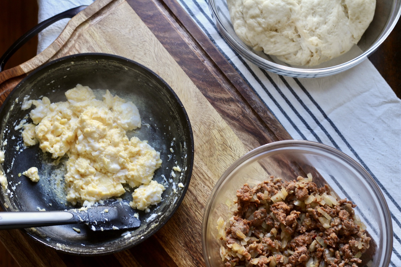 A bowl of kolache dough with scrambled eggs in a pan, cooked sausage in a bowl, and a cutting board on bottom.