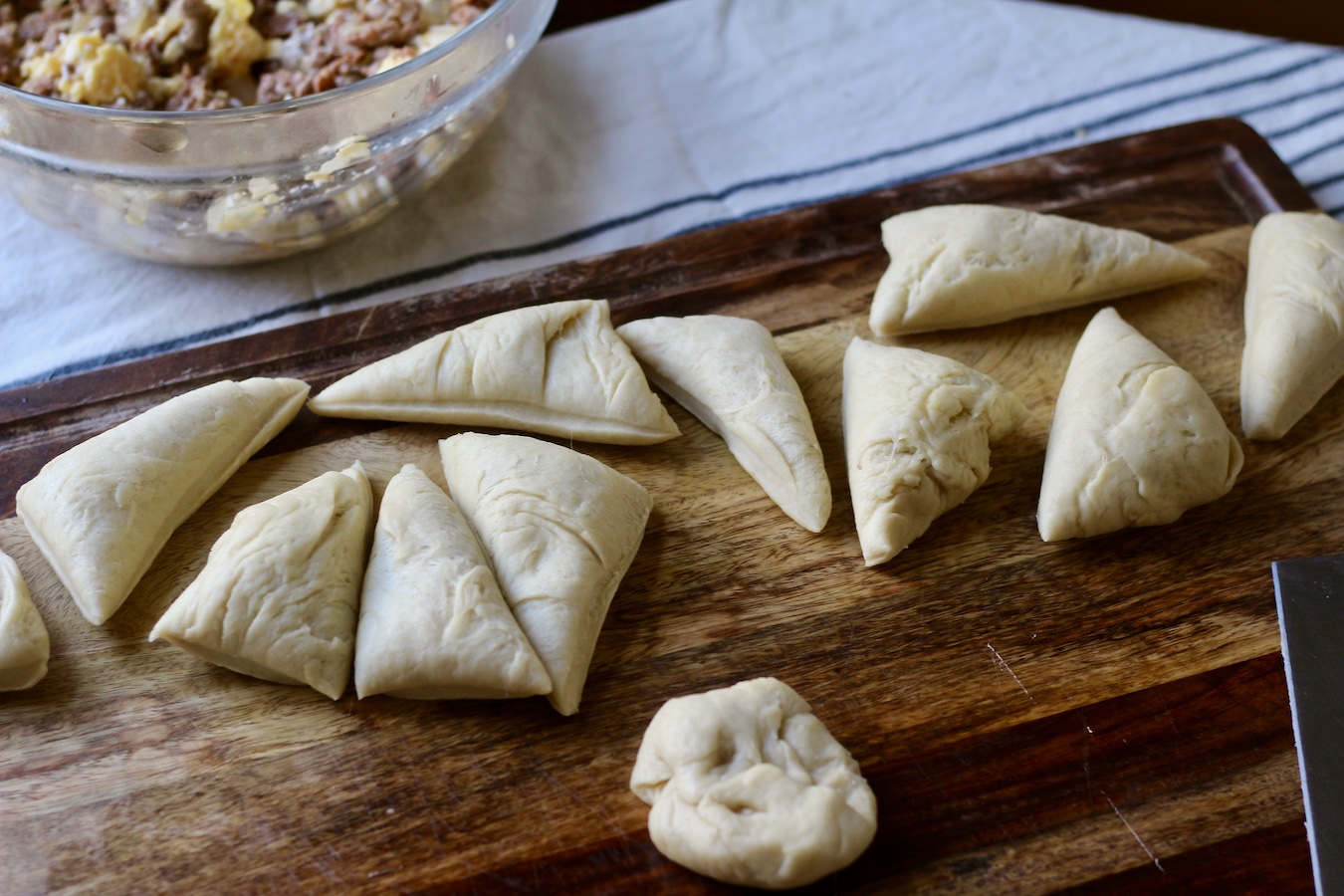 Kolache dough cut into pieces on a wooden cutting board with a bowl of sausage and egg scramble in the back.