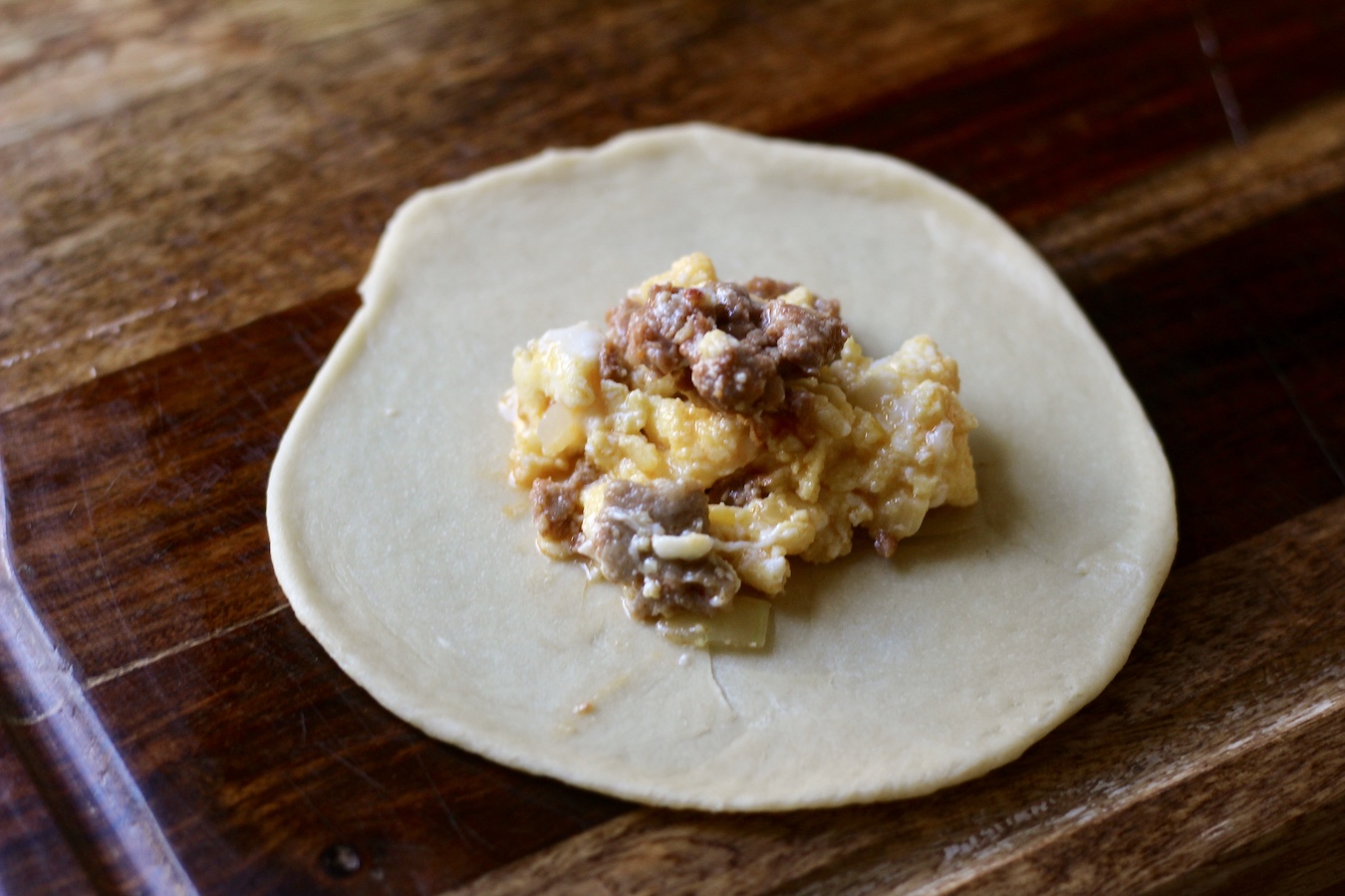 Round disk of koolache dough filled with sausage and egg scramble on a wooden cutting board.