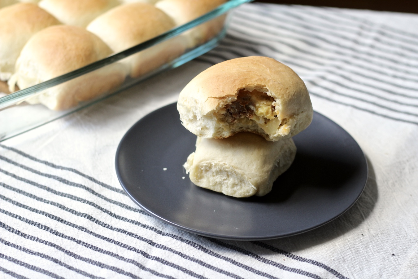 Two sausage and egg kolaches stacked on a blue plate with a platter of more kolaches in the back.