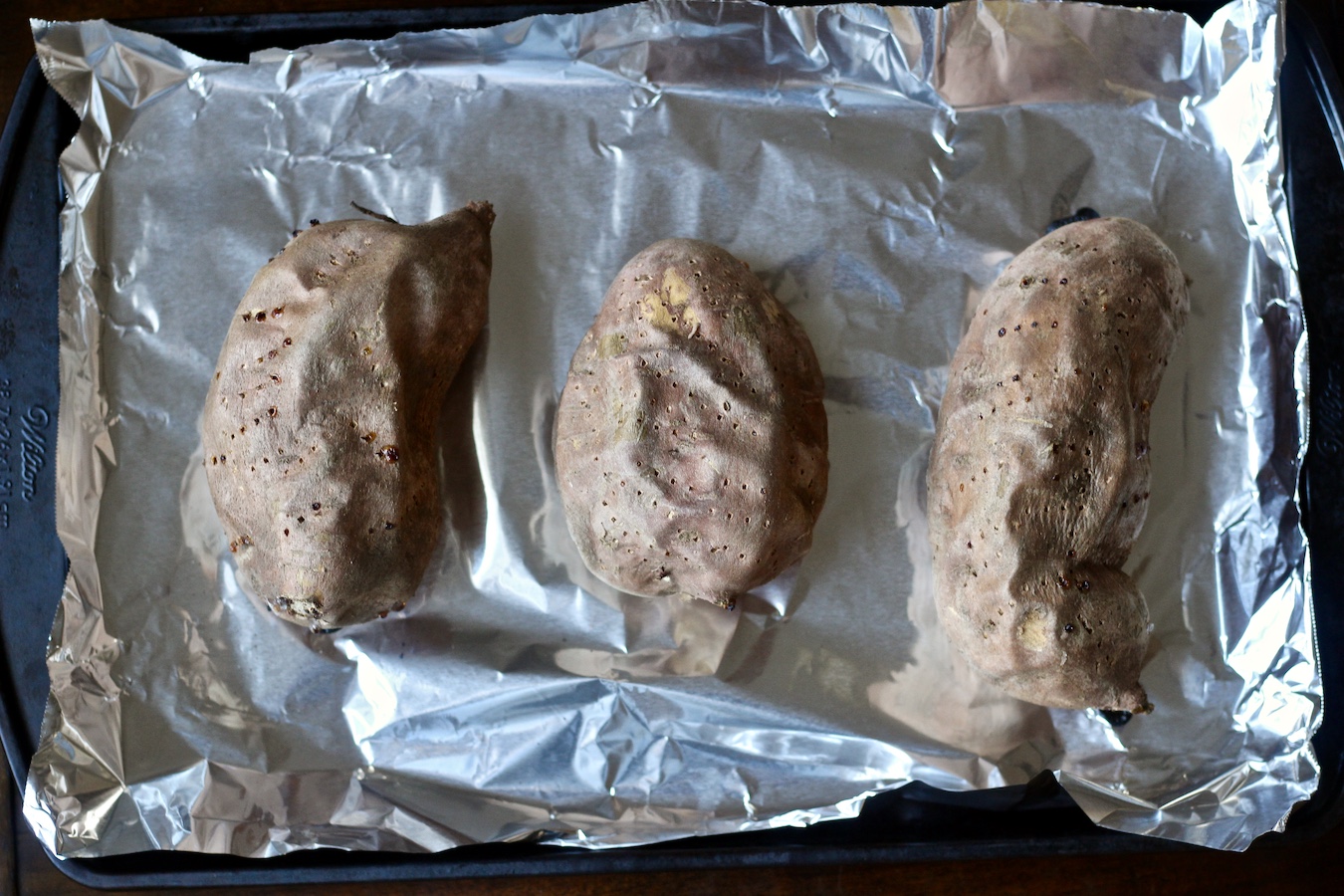 Three sweet potatoes on a baking sheet with aluminum foil.