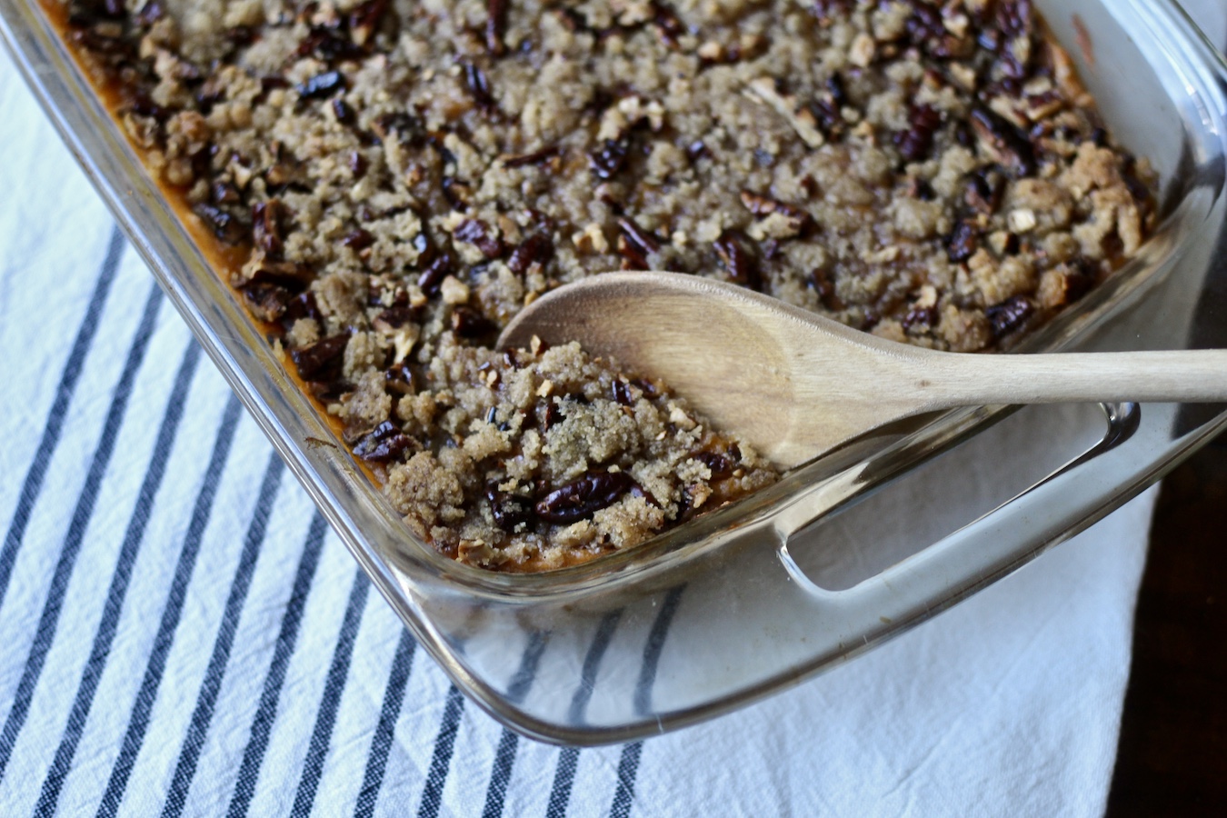 A wooden spoon inside the pyrex dish of sweet potato casserole on a white and blue kitchen towel.