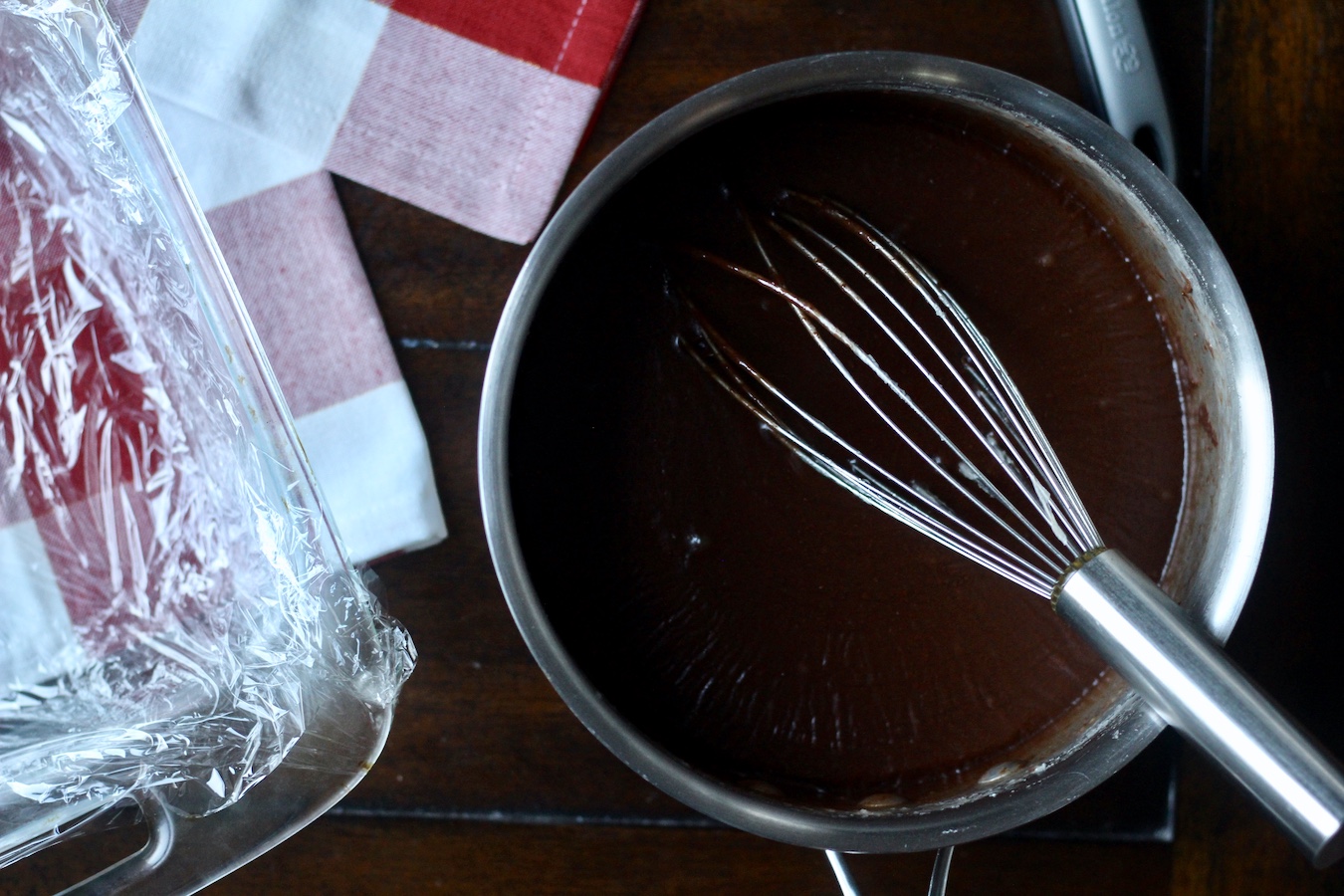 A silver pot of fantasy fudge on a wooden table with a prepared pan to the left.
