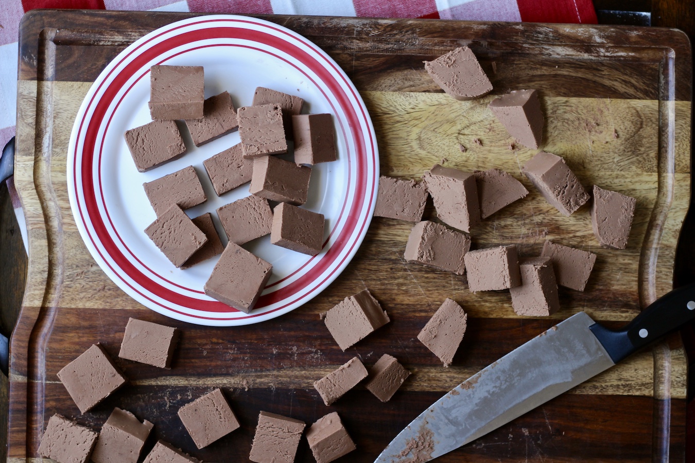 Traditional fudge cut into cubes on a white and red plate with more fudge on a wooden cutting board.