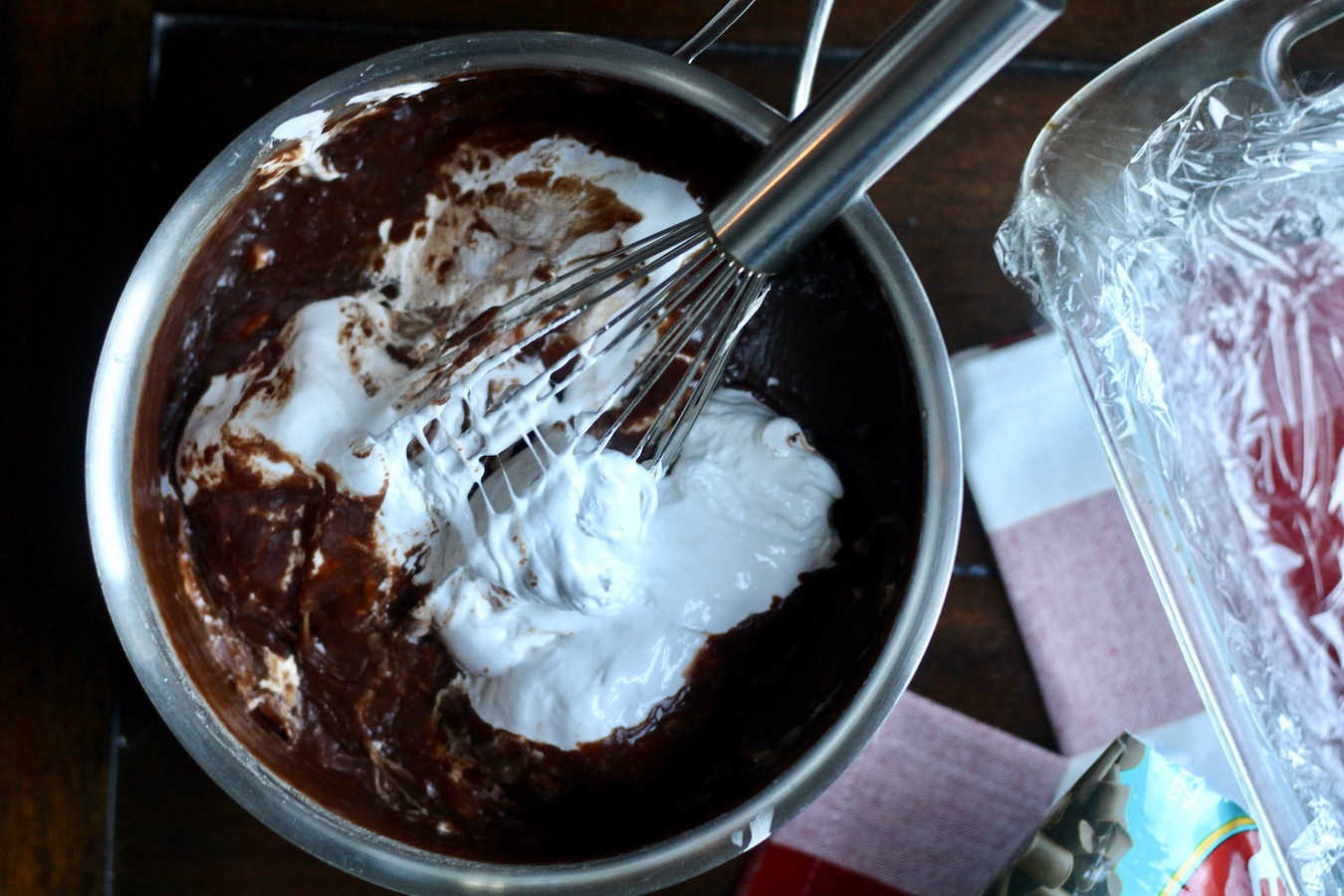 A silver sauce pan with fantasy fudge and marshmallow fluff being whipped with a whisk next to a glass pan on a red and white checkered towel.