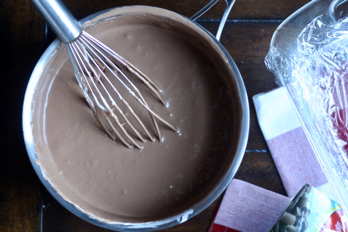 Smooth fantasy fudge in a sauce pan with a whisk next to a baking dish on a red and white checkered towel.