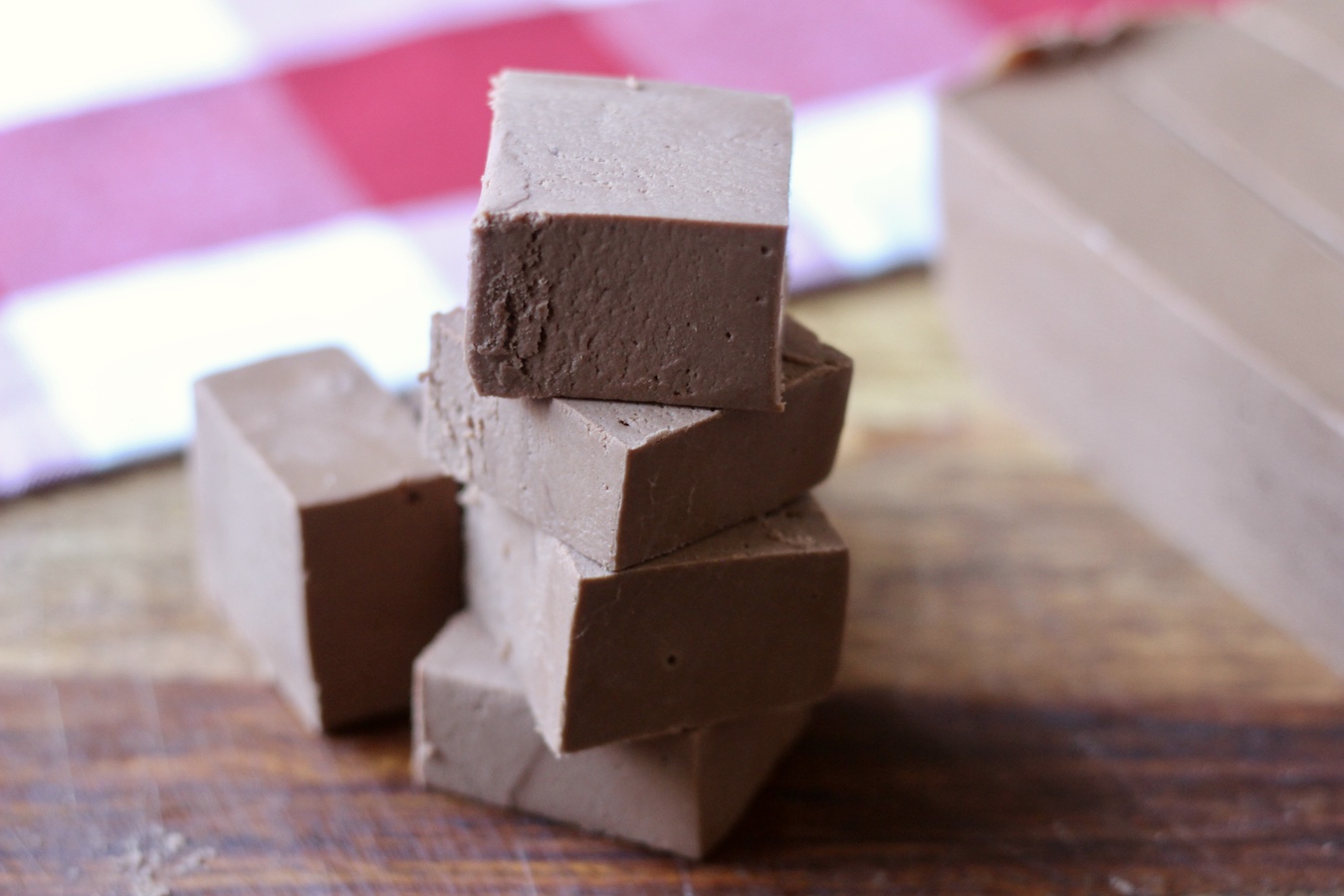 A small tower of fudge cubes stacked on a wooden cutting board with a erd and white checkered towel in the back