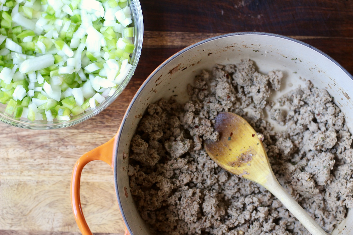 An orange pot with ground turkey and a small glass bowl of bell pepper and onion on a wooden cutting board.