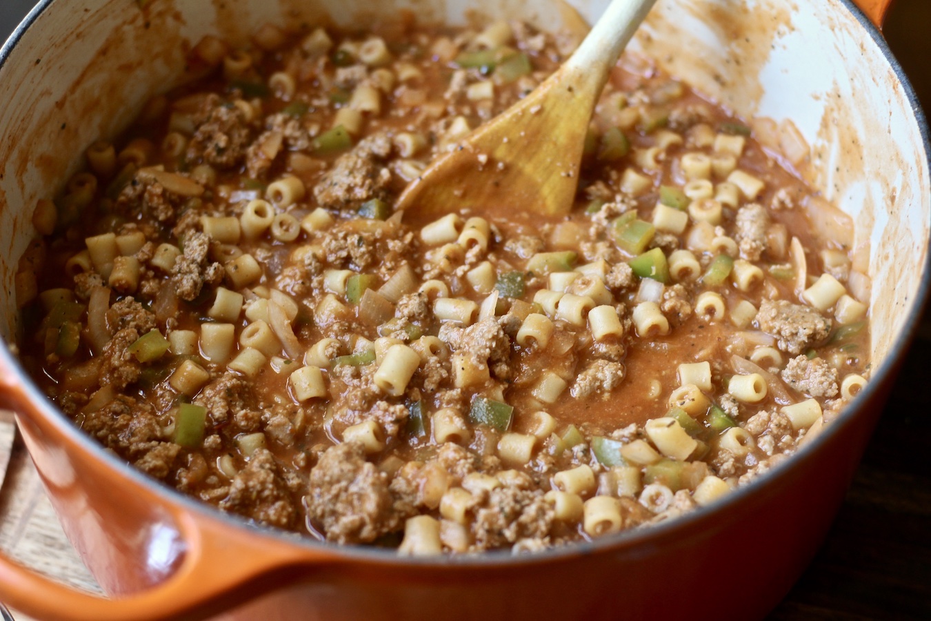 An orange pot of Turkey Mac with a wooden spoon on a wooden counter.