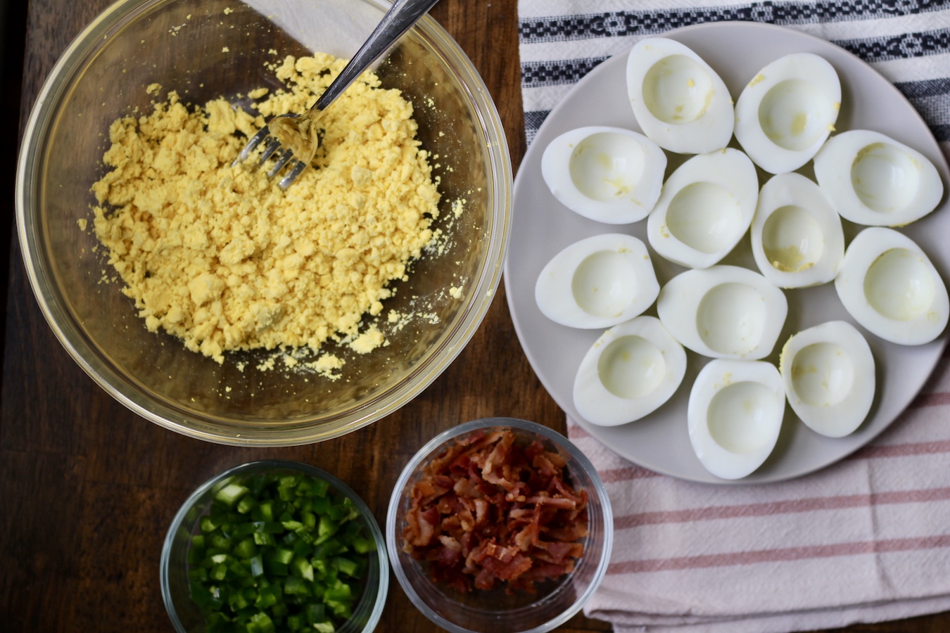 Ingredients fo Bacon Jalapeno Deviled Eggs in glass bowls on a wooden table with a blue and pink striped dish towel.