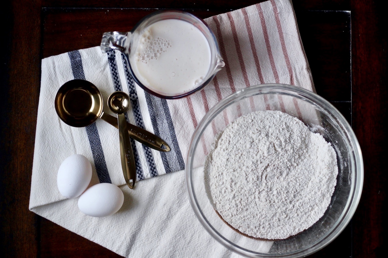 A glass bowl of flour, a liquid measuring cup of non-dairy buttermilk, two eggs, and two measuring spoons on a pink and blue striped dish towel on a wooden table.