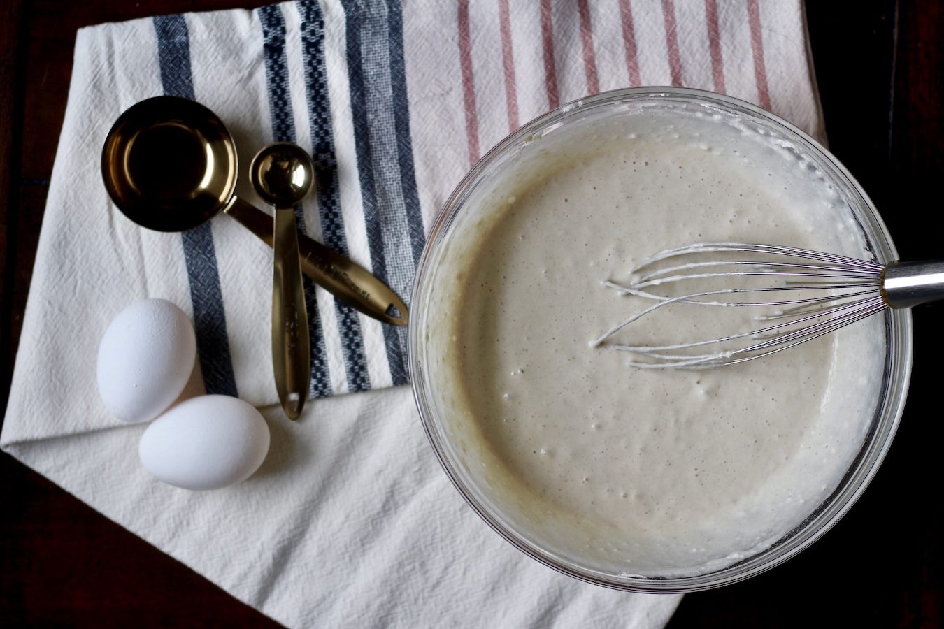 A glass bowl with buttermilk pancake batter and a silver whisk with two eggs, two copper measuring spoons to the left on a pink and blue striped dish towel on a wooden table.