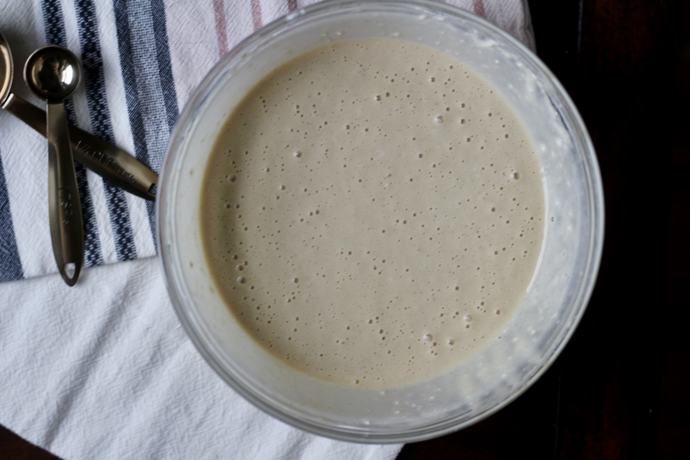 A glass bowl of buttermilk pancake batter with two copper measuring spoons to the left on a blue and pink striped dish towel on a wooden table.