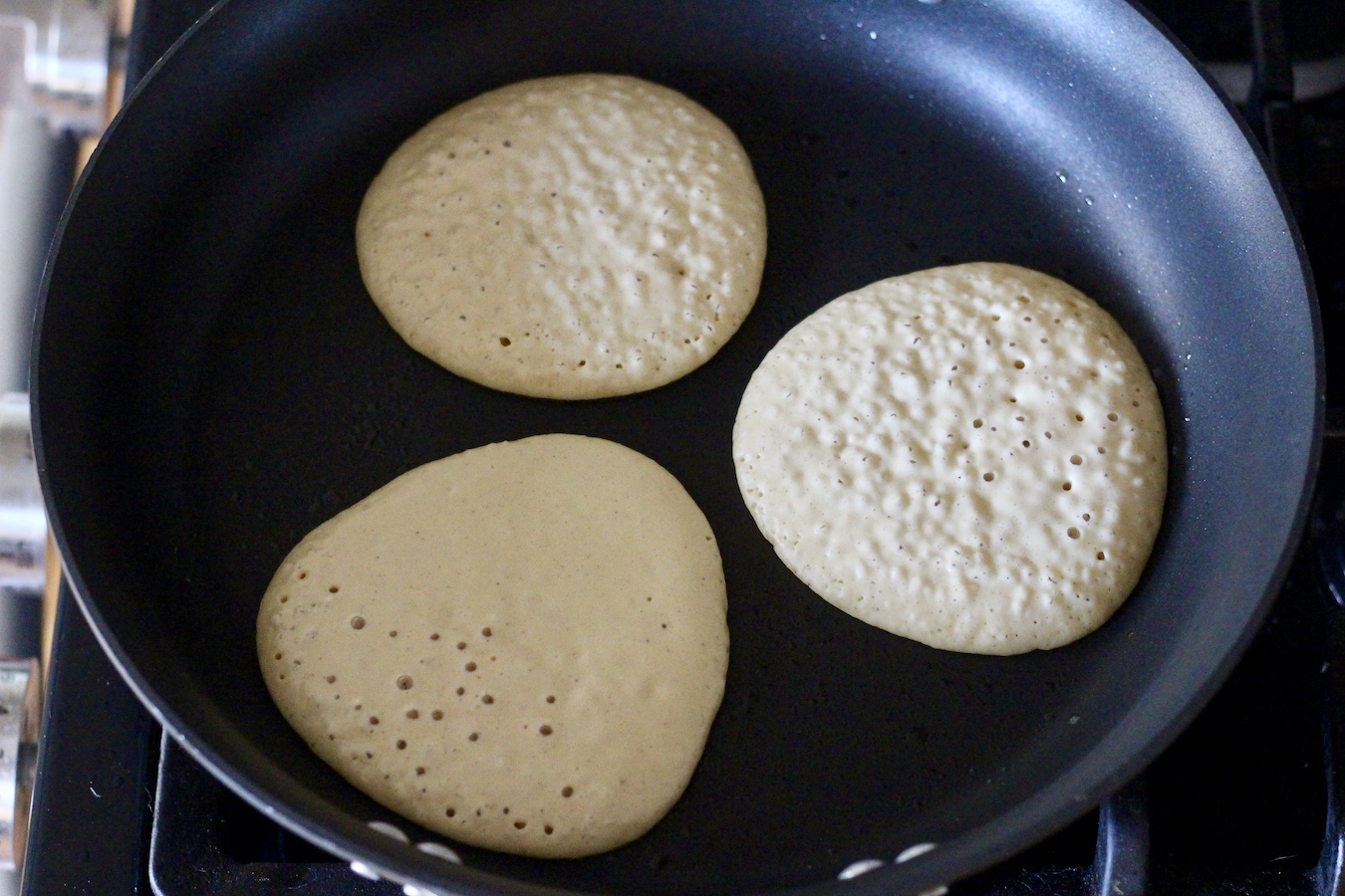 A black non-stick pan with three buttermilk pancakes cooking on a stovetop.