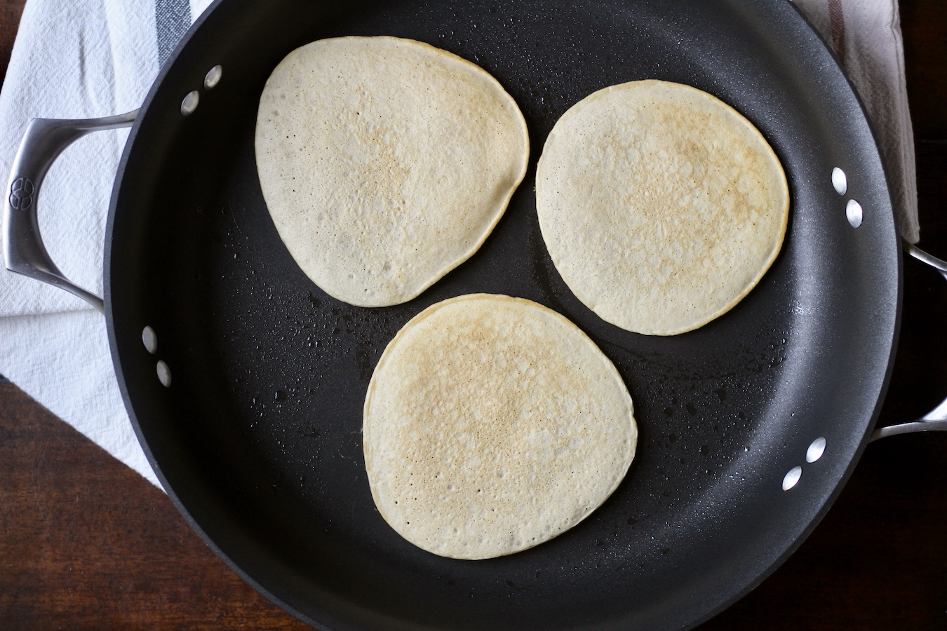 Three cooked buttermilk pancakes in a black non-stick skillet on white and blue striped towel on a wooden table.