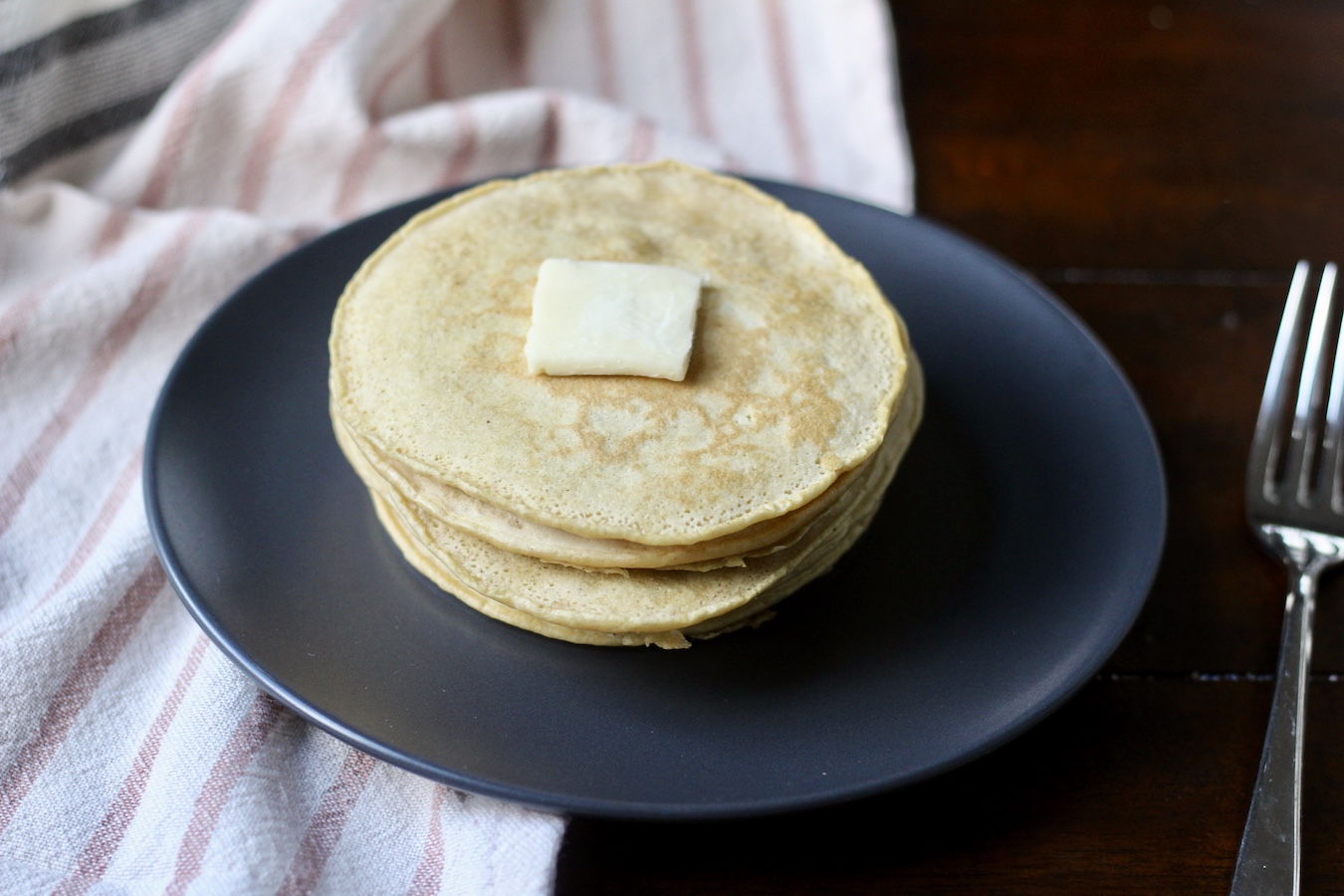 A stack of fresh buttermilk pancakes with a small square of vegan butter on top, on a blue plate on a white and pink striped dish towel wit a silver fork to the right on a wooden table.